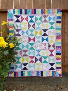 a colorful quilt next to yellow flowers in front of a wooden fence