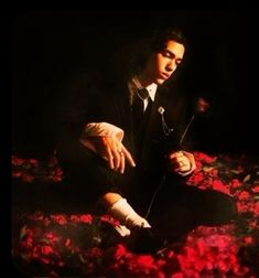 a man in a suit and tie sitting on the ground with red flowers around him