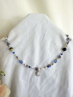 a necklace with beads and a crescent charm is displayed on a white cloth next to a flower