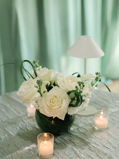 white flowers in a vase on a table next to candles and a light bulb with a green curtain behind it