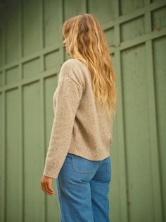 a woman with long blonde hair standing in front of a green wall wearing jeans and a sweater