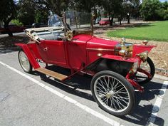 an old fashioned red car parked in a parking lot next to some grass and trees