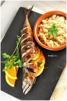 a fish and rice dish on a black plate with orange slices, garnished with parsley