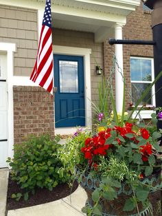 an american flag hanging from the side of a house next to some flowers and plants