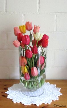 a glass vase filled with colorful tulips on top of a doily next to a brick wall