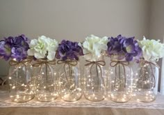 purple and white flowers in glass vases with string lights on the side, sitting on a lace tablecloth