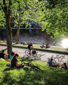 many people are sitting on the grass by the water and some have bikes in front of them