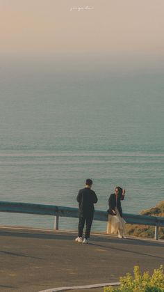 two people standing next to each other near the ocean with a kite flying in the sky