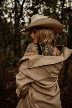 a woman wearing a hat and trench coat in the woods with tattoos on her back