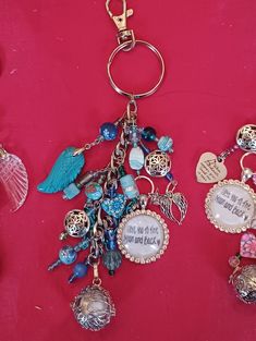 two key chains with charms attached to them on a red cloth covered surface, one has an angel charm and the other has a heart