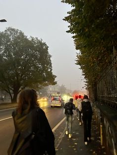 several people walking down the sidewalk in front of cars on a foggy day with trees lining the street