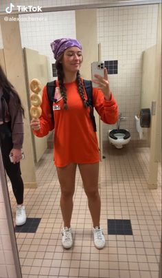 a woman in an orange shirt is holding two doughnuts and taking a selfie