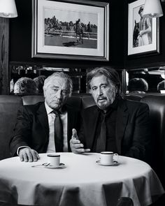 two men sitting at a table with cups and saucers in front of their faces