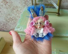 a hand holding a small doll with pink and blue feathers on it's head