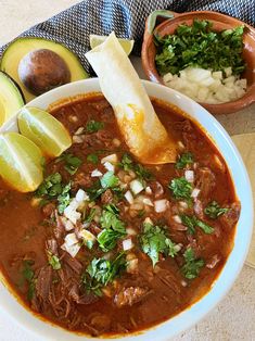 a bowl of chili soup with bread, avocado and cilantro on the side