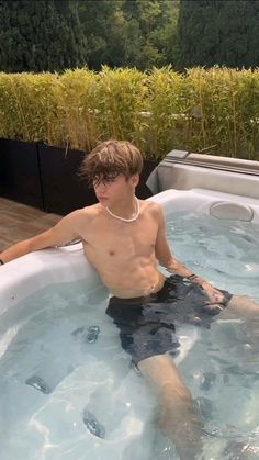 a young man sitting in a hot tub with water around his ankles and wearing a pearl necklace