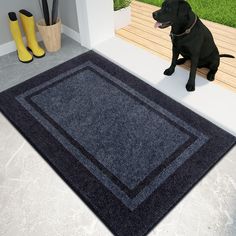 a black dog sitting on the floor next to a blue rug and yellow rubber boots