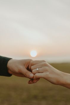 two people holding hands with the sun setting in the sky behind them and one person wearing a ring