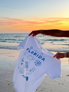 Our EXCLUSIVE Greetings From Florida Sunhoney T-shirt is perfect for beach lovers! This Comfort Colors t-shirt features a playful blue drawn design of popular Florida symbols like the sun, shell, mermaid, and more. Style this shirt over your fave bikini as your new go-to beach tee or style with biker shorts and sneakers for the cutest summer fit! Designed by us, handmade by us in Jacksonville Beach, FL Product is made to order: available for pick up/ships in 2-4 business days Content: 100% Cotto Custom Print T-shirt For Beach Vacation, Blue Beachwear T-shirt For Beach Party, White Graphic Tee For Beach Season, Fun Custom Print Tops For Beach Season, Blue Beach T-shirt With Screen Print, Fun White T-shirt For Beach Party, Blue Graphic Tee For Vacation, Blue Short Sleeve T-shirt For Beach Party, Short Sleeve T-shirt With White Print For Beach