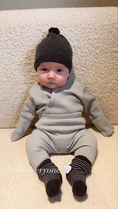 a baby sitting on the floor wearing a gray outfit and a black beanie hat