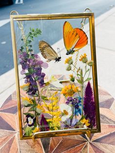 two butterflies sitting on top of a glass frame with flowers in front of it and a sidewalk behind them