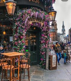 the outside of a restaurant with purple flowers growing on it's wall and people walking by