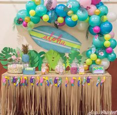 a table topped with lots of colorful balloons and desserts next to a surfboard