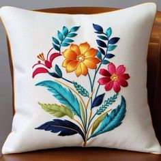 a white pillow with colorful flowers and leaves embroidered on the front, sitting on a wooden chair