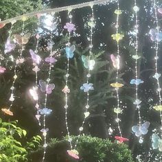 colorful glass butterflies hanging from a string in front of some bushes and trees with sunlight shining through the leaves