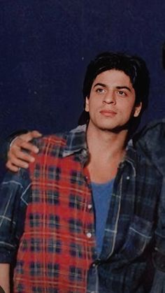 two young men standing next to each other in front of a blue wall and one is wearing a plaid shirt