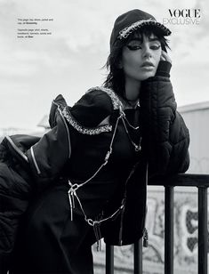 a woman leaning on a rail wearing a black coat and hat with chains around her neck