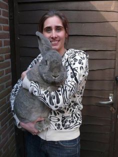 a woman holding a gray rabbit in her arms and smiling at the camera while standing next to a brick wall