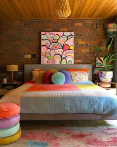 a bedroom with brick walls and colorful pillows on the bed, in front of a painting