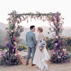 a bride and groom are standing under an arch with purple flowers on it at the top of a hill