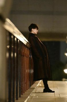 a person standing on a sidewalk next to a fence and railing with lights in the background