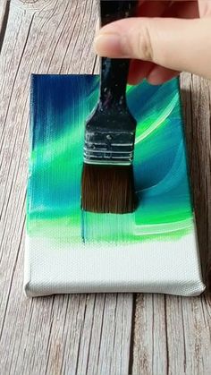 a hand holding a paint brush over a small painting on a piece of wood that has been painted green and blue