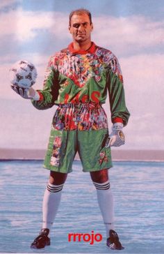 a man in a colorful outfit holding a soccer ball and wearing gloves with the ocean behind him