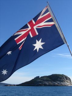 an australian flag flying in the wind on top of a boat with water and land in the background