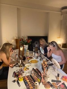 three women sitting at a table with makeup and other items on the table in front of them