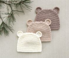 three crocheted bear hats sitting on top of a table next to pine branches