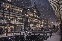 people walking in the snow at night near some buildings and benches with lights on them