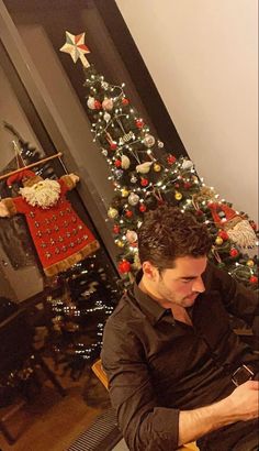 a man sitting in front of a christmas tree using his cell phone while looking at the screen