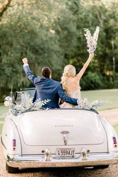 a bride and groom sitting in an old car with flowers on the roof, holding hands up