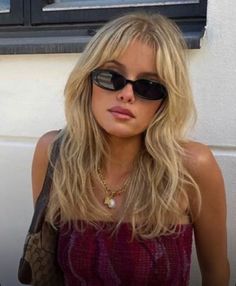 a woman with sunglasses and a handbag standing in front of a wall wearing a purple top