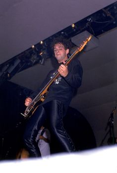 a man holding a guitar while standing on top of a stage with other people in the background