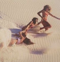 three women in bathing suits are on the sand with their surfboards and one is falling off her board