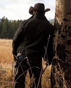 a man wearing a cowboy hat standing next to a tree in the middle of a field