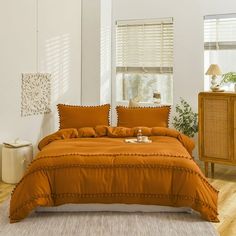 a bed with orange comforter and pillows in a white room next to a window