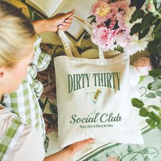 a woman is holding a tote bag next to some flowers and an open book