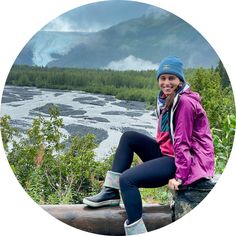 a woman sitting on top of a log in front of a river and mountain range